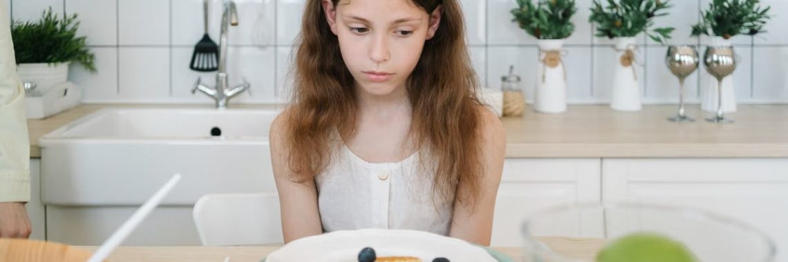 A young girl looks down unhappily at her breakfast pancakes with blueberries in a modern kitchen.